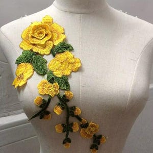 May include: A yellow embroidered floral applique featuring a large rose and smaller blossoms with green leaves. The applique is displayed on a mannequin, showcasing its potential use in fashion or craft projects.
