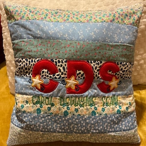 May include: A decorative pillow with a patchwork design. The pillow features horizontal stripes in various patterns, including floral, leopard print, and solid colors. The letters "CDS" are embroidered in red with star accents, and the name "CAROL DENAYNE SCAN" is embroidered below.