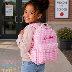 May include: A pink and white gingham patterned backpack featuring a ruffled trim along the front pocket and side panels. The name Zaria is embroidered in pink script on the front of the bag, which includes a top handle and adjustable shoulder straps.