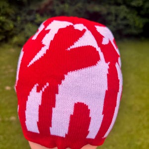 May include: A red and white knit beanie hat. The hat features a bold pattern of white shapes against a red background. The hat is made of a soft, knitted material and is designed to fit snugly on the head.
