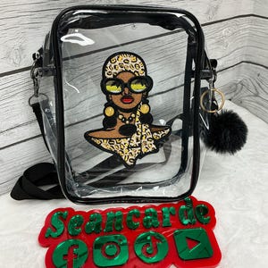 Può includere: Borsa a tracolla trasparente con cinturino e finiture nere. La borsa presenta una toppa ricamata di una donna con occhiali da sole e un foulard leopardato. Un cartello rosso e verde con icone di social media è di fronte alla borsa.