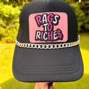 May include: Black trucker hat with a pink patch that says "Rags to Riches" in white and pink lettering. A silver chain is attached to the hat.