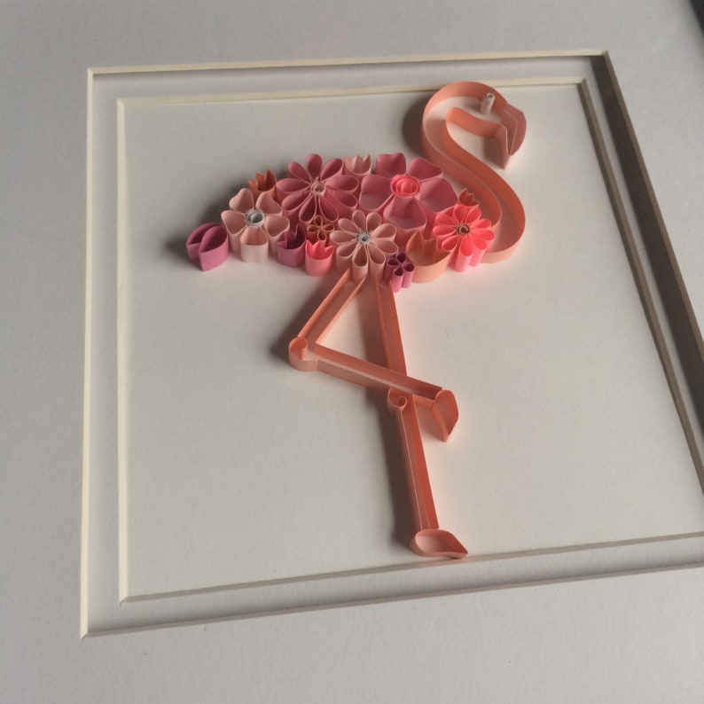 May include: A framed paper quilling art piece featuring a pink flamingo. The flamingo is crafted with rolled paper strips, with its body adorned with pink paper flowers. The artwork is set against a white background.