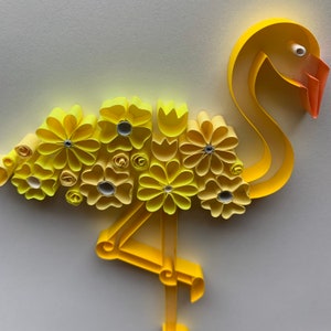 May include: A paper quilled flamingo sculpture in shades of yellow and orange. The flamingo's body is adorned with yellow paper flowers. The beak is orange, and the eye is white. The flamingo is standing on one leg.