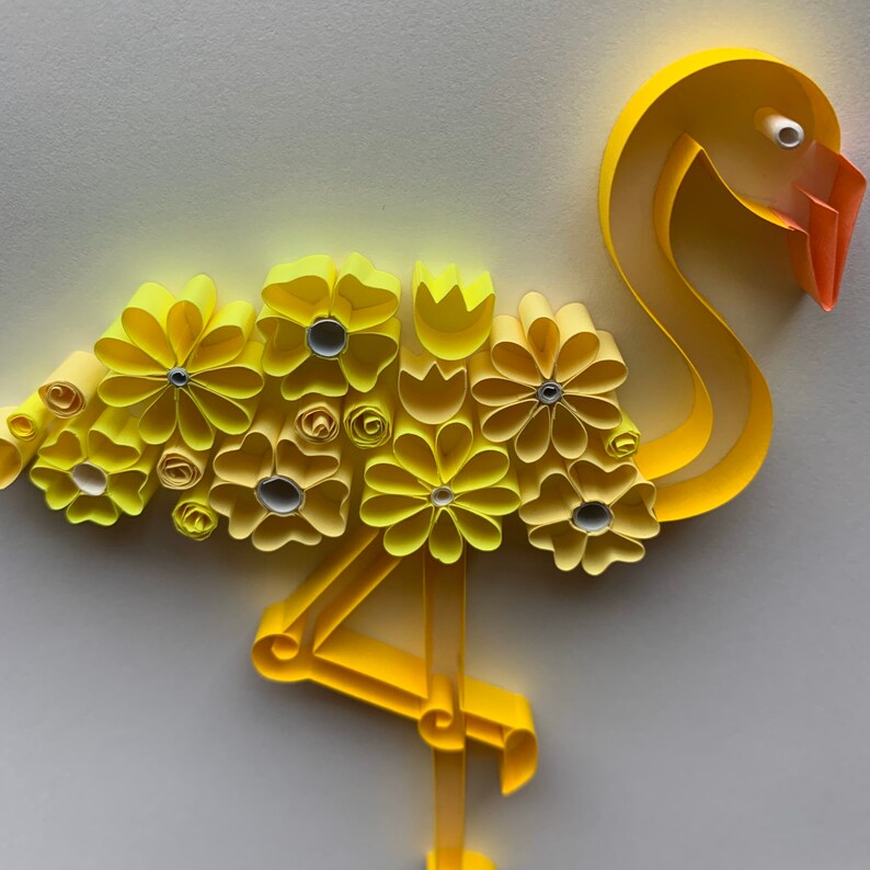 May include: A paper quilled flamingo sculpture in shades of yellow and orange. The flamingo's body is adorned with yellow paper flowers. The beak is orange, and the eye is white. The flamingo is standing on one leg.