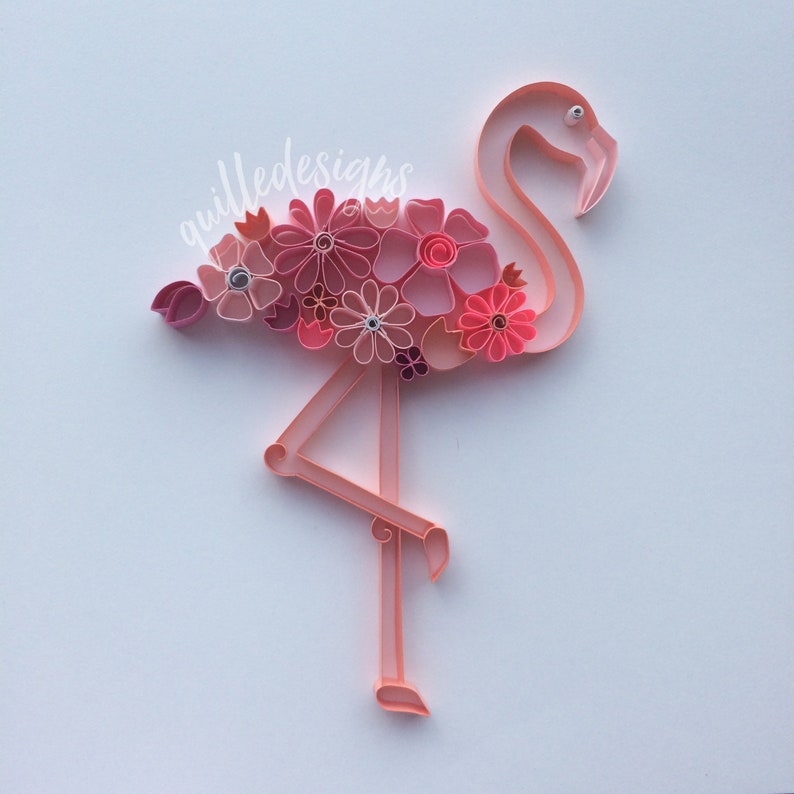 May include: A pink paper flamingo with a collection of pink and coral flowers on its back. The flamingo is standing on one leg. The text "quilleddesigns" is written in cursive on the upper left side of the image.