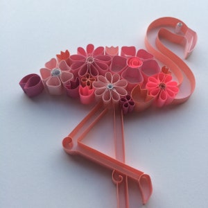 May include: A paper quilling art piece in the shape of a flamingo. The flamingo is made of pink paper strips and adorned with various pink paper flowers. The artwork is set against a white background, showcasing the flamingo's delicate details.