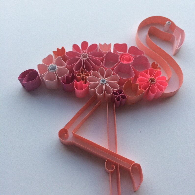 May include: A paper quilling art piece in the shape of a flamingo. The flamingo is made of pink paper strips and adorned with various pink paper flowers. The artwork is set against a white background, showcasing the flamingo's delicate details.