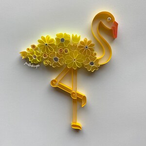 May include: A paper quilling art piece of a yellow flamingo decorated with yellow flowers and small silver accents. The flamingo has an orange beak and is set against a white background. The artist's signature, "gailledesigns", is visible.