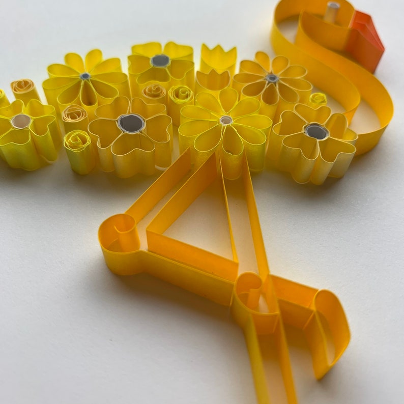 May include: A handmade paper quilling art piece featuring a bouquet of yellow flowers with silver centers. The flowers are arranged in a cluster, with a yellow ribbon-like element extending from the bouquet. The artwork is set against a white background.