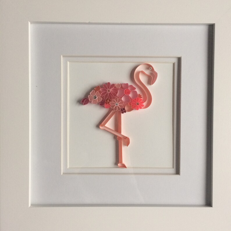 May include: A framed paper quilling art piece featuring a pink flamingo. The flamingo's body is adorned with pink paper flowers. The artwork is set against a white background within a white frame, creating a decorative wall art piece.