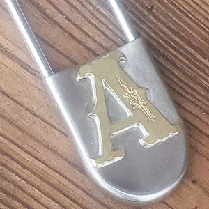 May include: A silver safety pin with a gold-colored letter 'A' and floral design on the front.