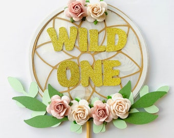 May include: A cake topper featuring a white and gold dreamcatcher with the words "WILD ONE" in gold glitter. The dreamcatcher is decorated with pink and white paper flowers and green leaves.