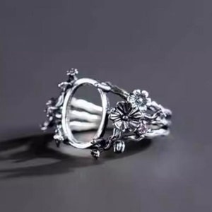 Può includere: Anello d'argento con una montatura ovale circondata da delicati dettagli floreali. L'anello presenta un design floreale con piccole pietre trasparenti.