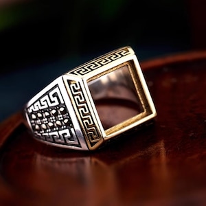 May include: A silver ring with a square, open-top setting. The ring is decorated with a black and gold geometric pattern.
