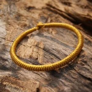 May include: A gold-colored braided bracelet made of cord. The bracelet is lying on a piece of weathered wood.