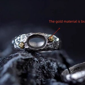 May include: A silver ring with a textured band and an oval-shaped setting. The ring has two small gold accents on the band. The gold material is brass.