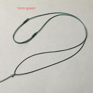 Puede incluir: Un collar verde oscuro con un cordón fino de 1 mm. El collar tiene un nudo ajustable y una pequeña cuenta redonda en el extremo. El texto "1mm green" es visible.