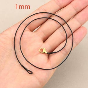Puede incluir: Un cordón de collar negro con un cierre dorado. El cordón tiene 1 mm de grosor. El collar se muestra en una mano.