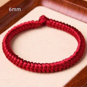 May include: A red braided cord bracelet with a 6mm width. The bracelet is made of a thick, woven cord and is a simple, elegant design.