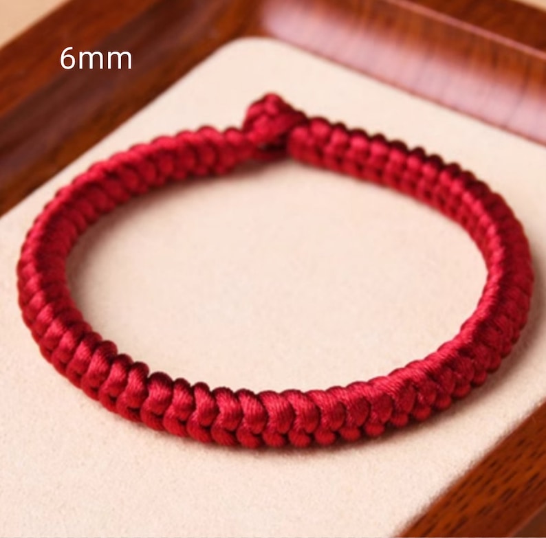 May include: A red braided cord bracelet with a 6mm width. The bracelet is made of a thick, woven cord and is a simple, elegant design.