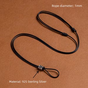 Puede incluir: Collar negro con un diámetro de cuerda de 1 mm, con una cuenta decorativa de plata de ley. El material es plata de ley 925. El collar se muestra sobre un fondo marrón.