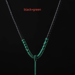 Könnte beinhalten: Eine schwarz-grüne Halskette mit geflochtenem Design. Die Halskette hat einen V-förmigen Anhänger mit einer langen, dünnen, grünen Quaste. Der Text "black+green" befindet sich in der oberen linken Ecke.
