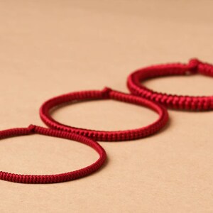 May include: Three red braided bracelets of varying sizes, arranged in a row on a beige surface.