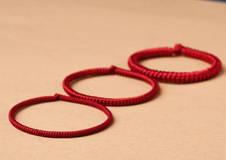 May include: Three red braided bracelets of varying sizes, arranged in a row on a beige surface.