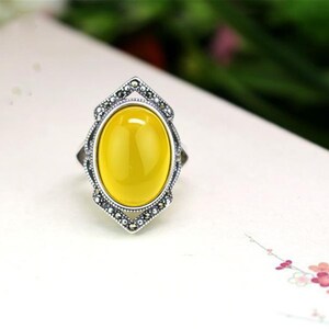 May include: A silver ring with a yellow oval gemstone set in a detailed silver frame. The frame is decorated with small black stones.