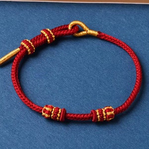 Peut inclure: Un bracelet en cordon rouge avec des accents dorés. Le bracelet comporte trois perles dorées aux motifs complexes.