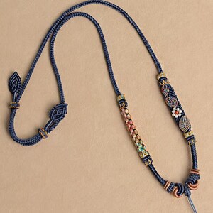 Puede incluir: Un collar de macramé azul marino con intrincados nudos y elementos decorativos. El collar presenta una sección central con cuentas coloridas y un diseño floral. Acentos dorados y detalles en forma de hojas realzan la elegancia. Fondo neutro.