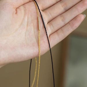 May include: A close-up of two delicate necklaces. One features a gold chain with a gold bar pendant, the other a black cord with a black bar pendant. The necklaces are displayed against a neutral background.