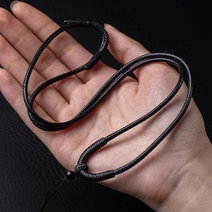 Peut inclure: Collier noir avec un cordon tressé et une petite perle ronde. Le collier est présenté sur une main, mettant en valeur son design simple et sa couleur sombre. Le cordon semble être fait d'un matériau durable.