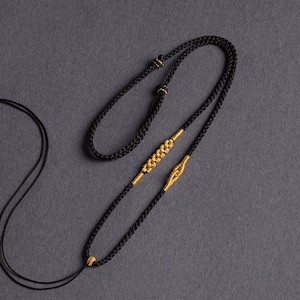 Peut inclure: Collier noir avec des accents dorés. Le collier présente un cordon noir tressé avec des éléments décoratifs dorés. Le design comprend une section centrale perlée et un détail en forme de fermoir, créant un look élégant et complexe.