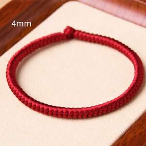 May include: A red braided bracelet with a 4mm width. The bracelet is made of a soft, woven material and is shown on a light brown surface.