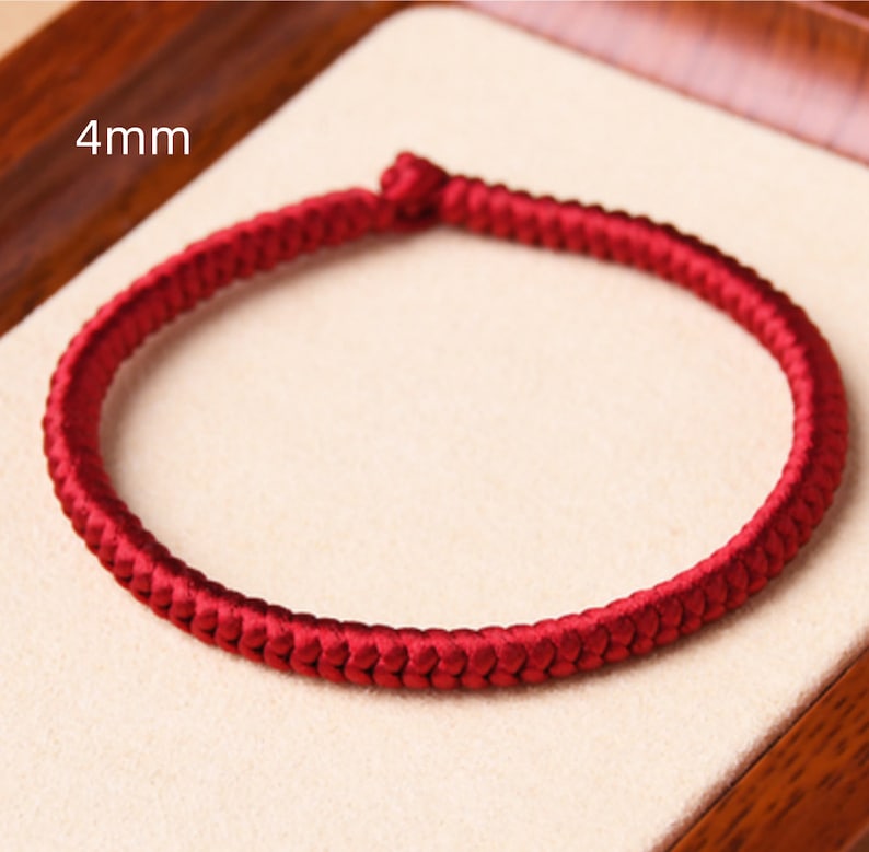 May include: A red braided bracelet with a 4mm width. The bracelet is made of a soft, woven material and is shown on a light brown surface.