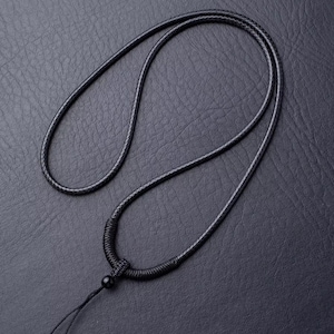 Puede incluir: Un collar de cuero negro con un diseño anudado y una pequeña cuenta negra. El collar se muestra sobre una superficie oscura y texturizada, mostrando su diseño simple pero elegante. El cordón mide aproximadamente 76 cm de largo.