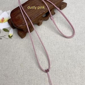 May include: A dusty pink necklace with a braided cord design. The necklace is displayed on a wooden stand. The text "dusty pink" is visible in the image.