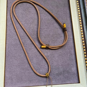 May include: A brown braided cord necklace with yellow detailing. The necklace is presented in a white and gold framed setting against a purple backdrop. The cord is knotted at the ends, featuring a simple, elegant design.