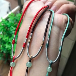 Puede incluir: Cuatro collares con colgantes de cuentas de jade. Los cordones son rojos, negros, turquesa y turquesa claro. Los cordones del collar están unidos a una pieza metálica plateada en forma de U. Las cuentas de jade son de color verde claro.