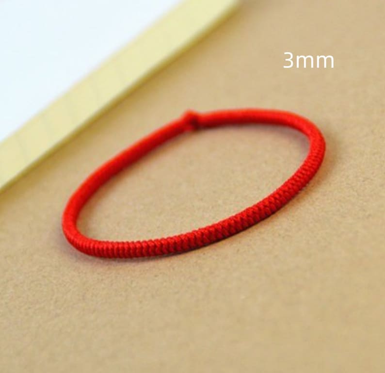 May include: A red string bracelet with a 3mm width. The bracelet is made of a thin, braided cord.