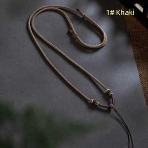Pode incluir: Colar cor cáqui com cordão ajustável. O colar apresenta pequenas contas verdes e douradas perto do pingente. O texto "1# Khaki" é visível. O colar é exibido em uma superfície cinza escuro.