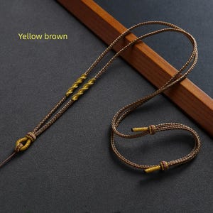 Puede incluir: Un collar amarillo-marrón con intrincados detalles tejidos y acentos dorados. El collar se muestra sobre una superficie oscura con un elemento de madera en el fondo. El texto "Yellow brown" es visible en la imagen.