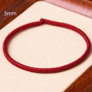 May include: A close-up of a red braided cord bracelet, 3mm in width, displayed on a light brown surface.