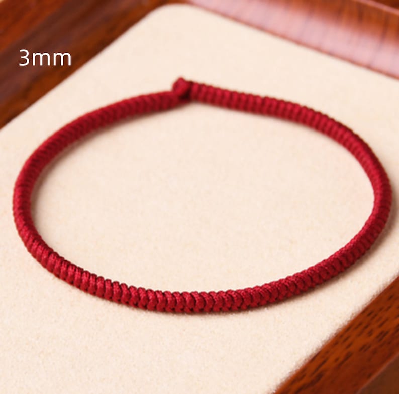 May include: A close-up of a red braided cord bracelet, 3mm in width, displayed on a light brown surface.
