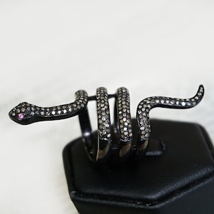 Puede incluir: Un anillo de serpiente de plata con diamantes negros y una piedra rosa para el ojo. La serpiente está enroscada y el anillo está en un expositor negro.