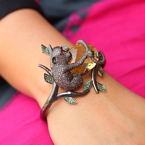 Op de afbeelding: Een zwarte en zilveren armband met een aap die een gele edelsteen vasthoudt. De aap is bedekt met kleine zwarte diamanten en de armband is versierd met groene bladeren.