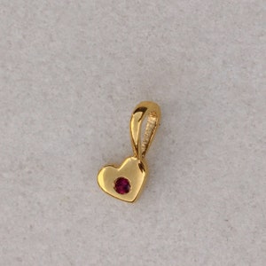 Puede incluir: Un colgante de corazón de oro con una pequeña piedra preciosa roja en el centro. El colgante tiene un bucle en la parte superior para sujetarlo a una cadena.