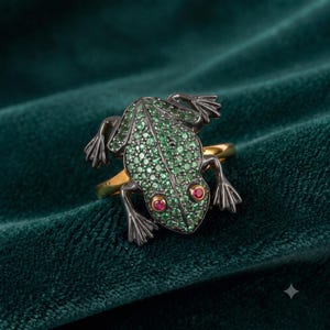 May include: A ring featuring a frog-shaped centerpiece crafted from dark metal. The frog's back is covered in small green stones, while its eyes are set with two round red stones. The ring band is a smooth, polished gold-toned metal.